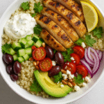 Grilled Mediterranean chicken bowl with quinoa, avocado, cherry tomatoes, Kalamata olives, cucumber, red onion, feta cheese, and tzatziki, served in a white bowl on a light background.