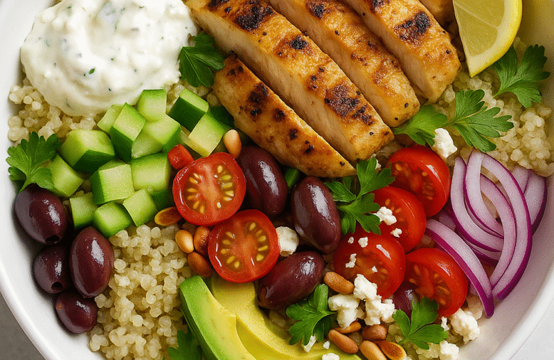 Grilled Mediterranean chicken bowl with quinoa, avocado, cherry tomatoes, Kalamata olives, cucumber, red onion, feta cheese, and tzatziki, served in a white bowl on a light background.